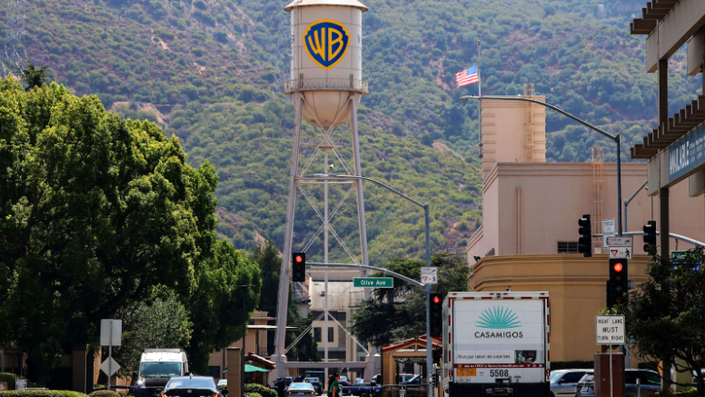 Warner Bros. water tower with WB logo in Burbank, California, surrounded by buildings, trees, and mountains in the background.
